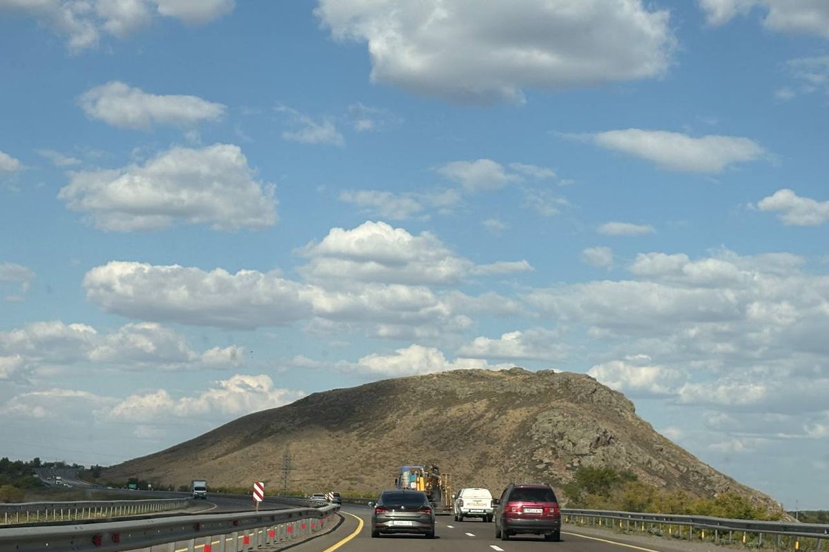 A two-lane road curving towards a bare hill under a cloudy sky
