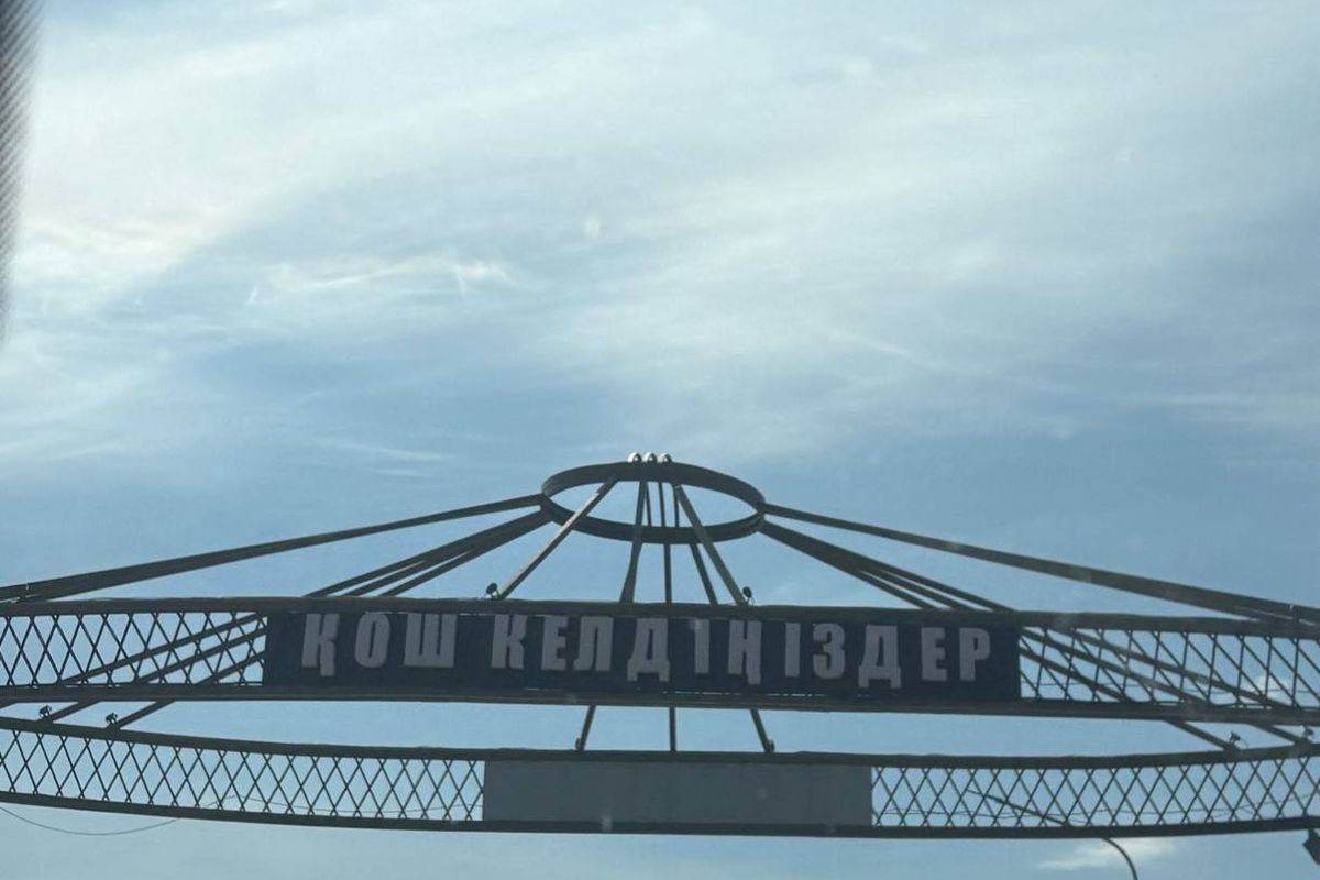 A steel arch shaped like a shanyrak over the road at a town entrance, with a welcome sign in Qazaq