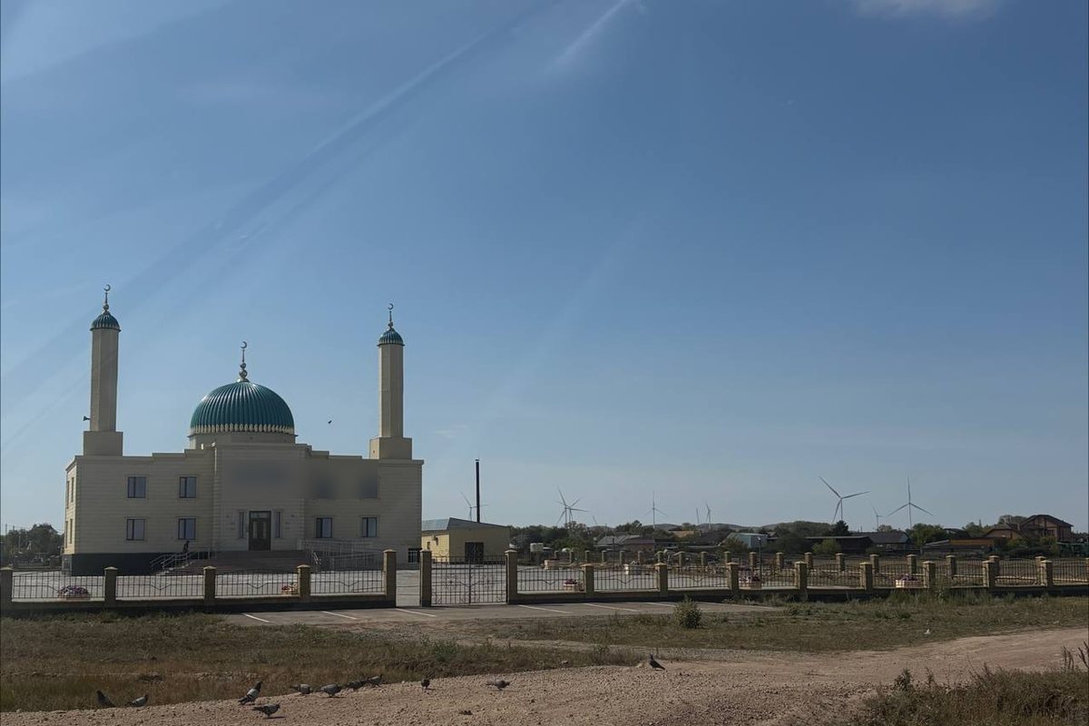 A small mosque with a green dome and two minarets behind a fence, wind turbines on the horizon